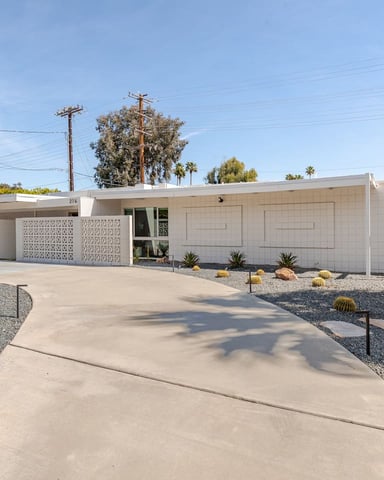 Mid-century modern white house with decorative garage doors, desert landscaping with cacti and gravel, concrete driveway, palm trees and power lines visible