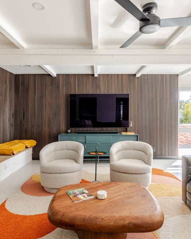 Modern living room with coffered ceiling, stone fireplace, two cream armchairs, wood coffee table, TV on fluted wall, and orange accents