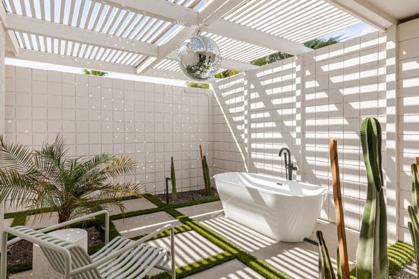 Outdoor bathroom with white tile walls, freestanding soaking tub, louvered pergola ceiling, disco ball, and desert plants including cacti and palms