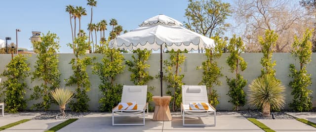 Poolside patio with two lounge chairs, white umbrella, wooden side table, and concrete wall with climbing vines