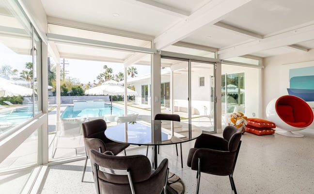 Modern sunroom with black chairs around glass table, red hanging chair, pool and palm trees visible through glass doors
